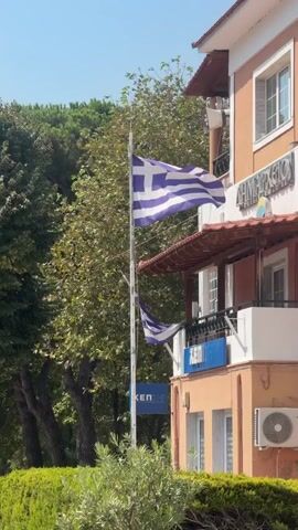 A Greek flag waves in the wind on a flagpole. Two smaller Greek flags are also visible. A building with orange and white walls stands to the right of the flagpole. Trees and bushes are visible in the background.