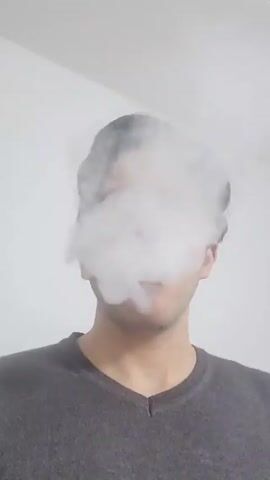 A person is seen from the chest up against a white wall. They are wearing a gray v-neck shirt. The person has hair that is visible at the top of their head and around their ears. A large cloud of white vapor or smoke surrounds their head, obscuring their face. The person extends two fingers to make a peace sign in front of the vapor.
