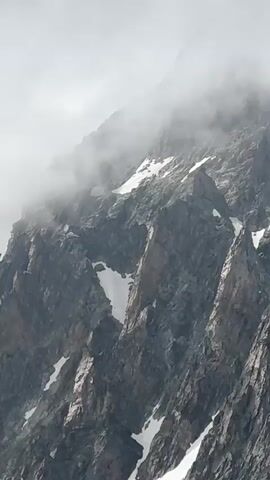 A video shows a panoramic view of a rugged, rocky mountain range. The peaks are partially obscured by swirling clouds and mist. Patches of snow are visible on the steep slopes, indicating a high altitude and a cold environment. The terrain is characterized by sharp, jagged rock formations and steep inclines, suggesting a challenging and remote landscape. The lighting is subdued, with the sky appearing overcast and gray. The camera pans slowly across the scene, revealing the vastness and imposing nature of the mountains. There are no people or man-made objects visible in the video.