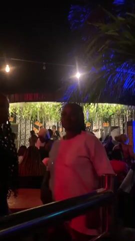 The video shows an outdoor gathering at night, possibly a party or an event. Several people are visible, some standing and others seated at tables. The environment is illuminated by string lights and colored lights, creating a festive atmosphere. There are palm trees and greenery in the background, suggesting a tropical or outdoor venue. Some individuals are facing the camera and smiling, while others are engaged in conversation or movement. The overall scene depicts a lively social event.