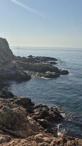 The video shows a rocky coastline with the sea on a clear day. The sun is shining brightly, casting shadows on the rocks. The water is a deep blue and appears to be calm with small waves lapping against the shore. There is a sailboat visible in the distance on the horizon. The foreground is dominated by rough, textured rocks. The sound of waves and birds can be heard throughout the clip.