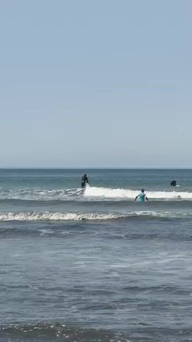 This media file captures a serene ocean scene with several individuals engaged in water activities. The primary subject is a person stand-up paddleboarding in the ocean, facing away from the camera. They are dressed in dark attire and are actively paddling. To the right and slightly further out at sea, another individual is visible on a paddleboard, also facing away. Further in the distance, a third person is seen on a paddleboard. The setting is a beach with gentle waves breaking on the shore, creating a foamy white crest. The water appears a mix of deep blue and lighter turquoise hues.

The time of day is likely daytime, indicated by the bright, clear sky and the sunlight reflecting off the water. The weather appears to be pleasant and fair, with a clear horizon. The location is identified as Costa da Caparica, Portugal, which is known for its expansive beaches and Atlantic waves, consistent with the visual cues in the media. No visible text is present in the file. The overall mood conveyed is one of peaceful recreation and enjoyment of the coastal environment.