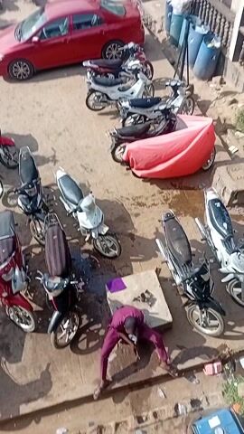 The video presents an elevated, wide-angle view of an outdoor area that appears to be a marketplace or commercial parking lot. The ground is predominantly paved and exposed, showing signs of use.

There is a dense cluster of parked motorcycles of various colors and styles, suggesting a popular mode of transport. Several cars are also parked among the motorcycles, including a red sedan prominently positioned in the center.

The backdrop of the scene is a two-story building complex featuring shops and businesses. The storefronts are adorned with large signs displaying brand names such as Tecno, Infinix, and Itel, indicating a focus on technology and mobile devices. There are people standing and walking on the second-story walkway of the building, as well as on the ground level near the shops.

The environment suggests a bustling commercial area in a daytime setting. The overall scene implies a busy and active local marketplace.
