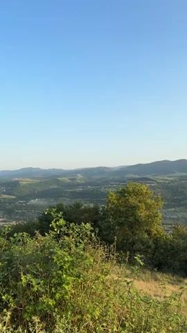 A panoramic view of a valley with rolling hills and a clear blue sky. The foreground features lush green vegetation and dry grass. In the midground, trees are scattered across the landscape, and a small town or village is visible nestled in the valley. The background consists of more hills that fade into the distance. The lighting suggests it is daytime, possibly late afternoon, given the soft, warm light on the hills.