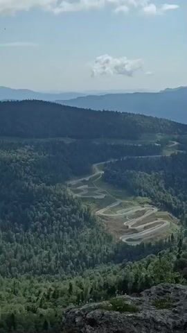 The video shows a panoramic view of a mountainous landscape with a winding road carved into the hillside. The road has many switchbacks and appears to be under construction or recently completed, with exposed earth and gravel in some sections. The surrounding mountains are covered in dense green forest, and several layers of distant, blue-hued mountains are visible on the horizon. The sky is partly cloudy with dramatic cumulus clouds. In the foreground, a rocky outcrop with some sparse vegetation is visible. The camera pans slowly from left to right, revealing more of the vastness of the landscape. There are no people or vehicles visible in the video. The overall impression is one of a remote and rugged natural environment, with human intervention in the form of the road being a prominent feature.