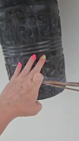 A person's hand with neon pink and green nail polish is holding a bundle of incense sticks. The incense sticks are lit and are creating smoke. The hand is positioned in front of a dark, cylindrical object with embossed lettering, which appears to be a prayer wheel. The prayer wheel is mounted on a metal bracket against a white wall. The background shows another similar prayer wheel and a shelf.