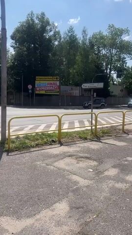 A video captured from street level shows a pedestrian crossing at an intersection on a sunny day. A white van is seen crossing the intersection. A traffic light is visible, indicating that traffic is currently stopped. The camera angle is from behind a yellow barrier, with a sidewalk in the foreground. The background features trees and buildings. A billboard with Polish text is also visible.