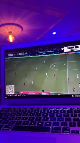 A person's hand, partially visible on the left side of the frame, holds a laptop computer which displays a soccer match. The game is in progress, with players on a green field. The score appears to be ESP 1 - 0 KSA. The words FIFA WORLD CUP 2026 are visible on the screen. The room is lit with purple and blue lights, casting shadows on the ceiling. The laptop is a MacBook Air.