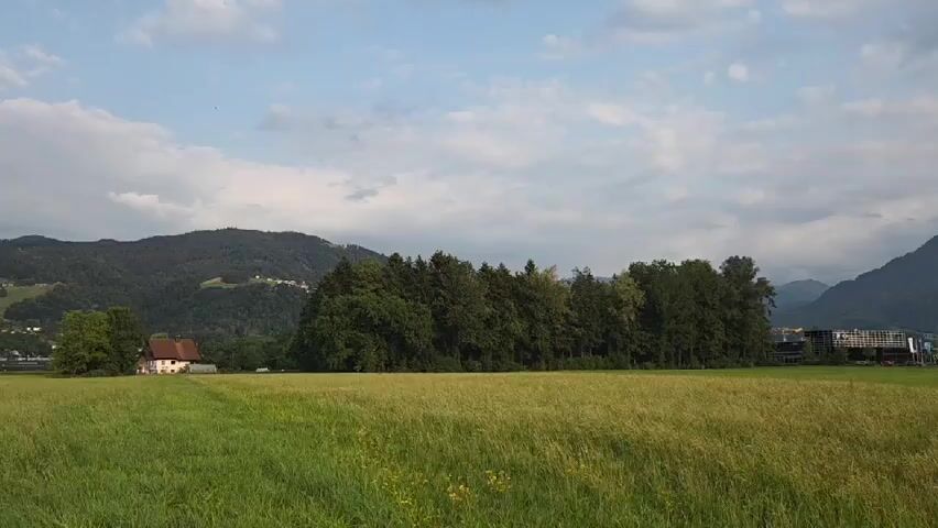 A drone shot pans slowly across a vast, green field of grass under a cloudy sky. In the distance, rolling hills covered in trees and dotted with houses form a picturesque backdrop. A lone house with a brown roof stands to the left of the frame, near a small cluster of trees. The camera continues to move, revealing more of the green landscape and the distant mountains.