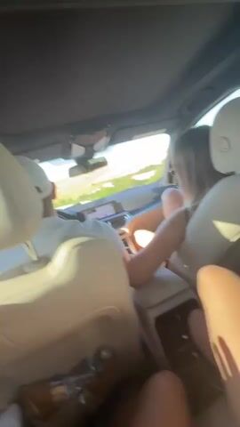 The video shows the interior of a car from a backseat perspective. There are three people visible. In the passenger seat in front, a person with blonde hair is visible from the back. The driver is partially visible on the left side of the screen. The legs of a person in the backseat are visible in the foreground on the right side of the screen. The car appears to be moving, with scenery visible through the windows.
