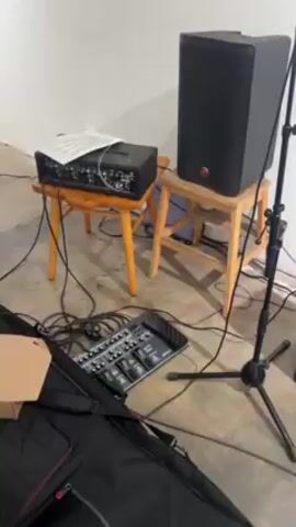 The video begins with a view of music equipment including a mixer, speaker, and guitar case. The camera then moves to show an art gallery with several framed artworks on white walls. A yellow fan stands in the corner of the room. A man with curly brown hair, wearing a white shirt and a black button-down shirt, turns to face the camera in front of one of the artworks. He has glasses and a light beard. The camera then zooms in on the artwork behind him, which depicts a pickup truck in front of a red building with signage. The man is then partially visible in the foreground.