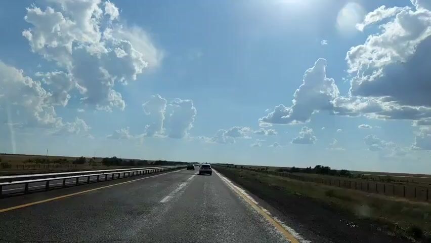The video shows a first-person perspective of driving on a highway on a sunny day. The sky is blue with scattered white clouds. The sun is visible in the upper right corner of the frame, and its rays are casting a glare on the scene. The highway is asphalt with yellow and white lane markings. There are guardrails on the left side of the road and dry, grassy fields with some scattered bushes and trees on both sides. Two cars are visible on the highway ahead, also traveling in the same direction. The car in front of the viewer is a dark sedan, and the car further ahead is a lighter-colored SUV. The overall impression is one of travel through an open, rural landscape.