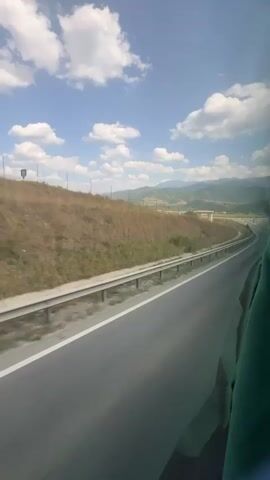 The video shows a view from inside a moving vehicle, likely a bus, looking out a window at a highway. The road curves to the right, bordered by a metal guardrail. Beyond the guardrail is a dry, grassy embankment with scattered green bushes. In the distance, rolling hills and mountains are visible under a bright blue sky dotted with white, fluffy clouds. The time stamp indicates the video is only a few seconds long, capturing a moment of travel through a rural or semi-rural landscape. The right side of the frame shows a dark, green fabric, possibly part of the vehicle's interior or a person's clothing.