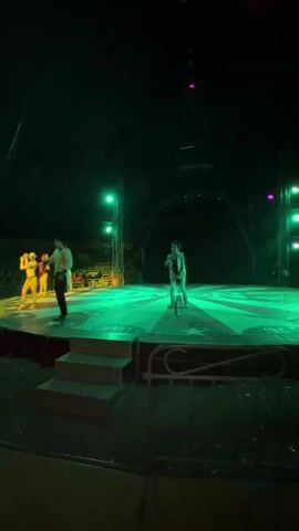 The video shows a circus performance in progress.  A young performer is riding a unicycle in the center of the stage. The stage is round with a red and white radiating design. A man in a white shirt and black pants is walking on the edge of the stage. A woman in a sparkly outfit is gesturing with her hands, also at the edge of the stage. Stairs lead up to the stage. Lighting rigs and equipment hang from the tent ceiling. The lights change from red to green to blue. Seating can be seen in the background.