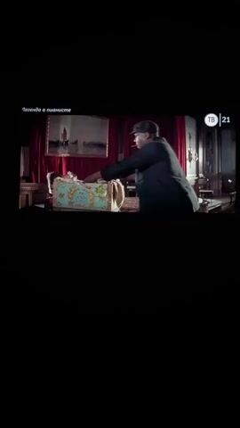 A man in a dark hat and jacket is seen bending over a large decorative trunk. He appears to be placing something inside. He then lifts a baby, holding it up and looking at it. The background is dimly lit, with red curtains and a painting on the wall. A TV channel logo is visible in the upper right corner.