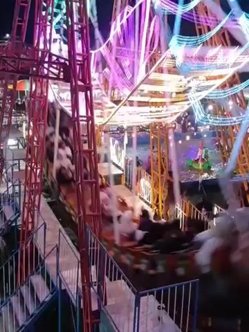 The video shows a spinning amusement park ride at night. The ride is made of a red metal structure with colorful lights. Several people are seated in carriages that are rotating around the center of the ride. The carriages appear to be moving rapidly, causing a blur of motion. The overall atmosphere is festive and exciting.
