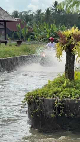 A man in a black and white shirt, black shorts, and a red helmet rides a red ATV through a shallow canal of water, splashing water around him. The man is facing forward, and the ATV is moving from left to right across the frame. In the background, there is a small brown building with a red roof, green grass, and trees. Another ATV is visible in the distance. The sky is partly cloudy.
