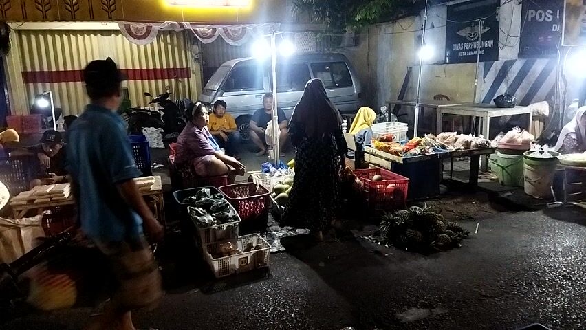 A nighttime street market scene is depicted. In the foreground, a person walks away from the camera. To their left, a man is seated at a wooden stall piled with goods, possibly produce. To the right, several other individuals are visible around various market stalls. One stall displays an assortment of fruits, including what appear to be durians. Another stall has more produce and possibly packaged items. In the background, a silver van is parked, and several lights illuminate the market area. The overall impression is a busy, informal marketplace.