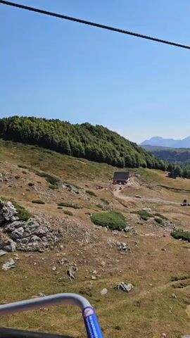 The video shows a mountain landscape with a building and several animals. A ski lift cable is visible in the foreground, suggesting the footage is taken from a moving ski lift. The terrain is rocky and covered with dry grass and scattered bushes. A forest of green trees covers the slope of a mountain in the background. A building, possibly a lodge or cabin, is situated on the mountainside, with a red tractor parked outside and several cars nearby. A group of horses and cows are gathered in a field near the building. The sky is clear and blue.