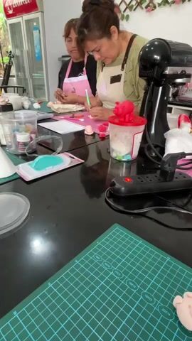 A group of people, mostly women, are gathered around a large black table, seemingly participating in a cake decorating class. Two women in the foreground are actively engaged in working with pink fondant or modeling paste, using small tools. One woman, wearing an apron, is carefully shaping a piece of pink material with a green tool. Beside her is a red container with a pig figurine on top, filled with white material and colorful objects. Another woman, also wearing an apron, is seated to her left, focused on her task with pink fondant. In the background, a man is visible, also working with materials on the table. Various cake decorating tools and supplies, including piping bags, small containers of colored powder, a white cone, and a green cutting mat, are spread across the table. A black stand mixer is positioned to the right of the women. The environment appears to be a brightly lit workshop or kitchen, with a refrigerator visible in the background and greenery on the wall. The overall scene suggests a hands-on learning or creative activity related to baking and confectionery.