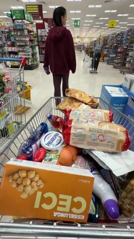 Here is a detailed description of the video:

The video shows a shopping cart filled with various groceries in a brightly lit supermarket. The camera is positioned inside the shopping cart, looking forward as it is pushed through the store. Dominating the foreground are items such as a box of Esselunga brand chickpeas, a package of Philadelphia cream cheese, loaves of Mulino Bianco bread, bottles of cleaning products, and bags of chips. The cart also contains other miscellaneous items, like a sponge and some packaged olives, creating a varied assortment of goods.

A person wearing a maroon hoodie and pants walks ahead of the cart, pushing it through the aisle. The supermarket is well-stocked with various products on shelves lining both sides of the aisle. Additional shopping carts and displays of merchandise, such as umbrellas, can be seen in the background, further indicating a busy shopping environment. The floor is a light-colored tile, and ceiling lights provide ample illumination to the entire scene.