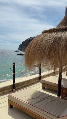 The video shows a beach resort with two lounge chairs under a thatched umbrella overlooking a body of water. The water is a clear turquoise color and there are boats in the distance. There is a rocky coastline with some buildings on it. The sky is blue with some white clouds. There are a few people in the water, and one person standing on the shore. The lounge chairs are made of wood with beige cushions and red trim. The umbrella is made of straw.