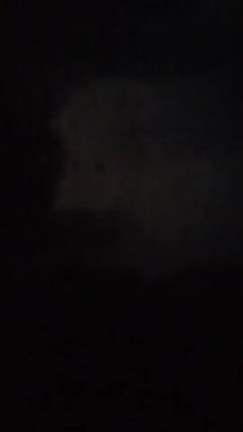 The video begins with a dark, blurry shot of what appears to be water droplets on a windowpane, with faint, indistinct shapes visible through the glass. The scene is very dark, and the shapes are difficult to discern. The video then cuts to complete blackness for several seconds before showing another glimpse of the blurry, dark scene through the window. The shot is unstable and shaky, suggesting it may have been recorded by hand. No people or specific objects are clearly identifiable. The environment appears to be indoors looking out, possibly at night or during a storm. The overall impression is one of obscurity and darkness.