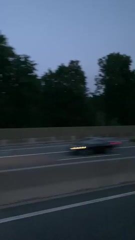 The video shows a blurred, side-view of a highway at dusk or dawn. The sky is a deep, muted blue. On the left side of the frame, dark trees are visible, some appearing to sway due to motion blur. A beige barrier runs along the highway. The highway itself is dark asphalt with white lines indicating lanes. In the distance, a dark, tree-covered hillside is visible on the right. There are no people or other vehicles clearly discernible in the video, only the impression of movement. The overall impression is of a car traveling along a road through a wooded area as light conditions are low.