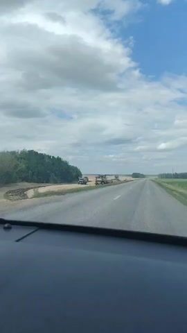 The video shows a view from inside a vehicle, looking out at a rural landscape. A paved road stretches into the distance, with a field on the right and a tree line on the left. Several agricultural vehicles are visible, including a combine harvester, trucks, and possibly tractors. One truck is moving along a dirt path beside the road, appearing to be loaded with material. The sky is partly cloudy with blue sky visible between the clouds. The weather appears to be fair. The overall scene suggests agricultural activity or road construction in a rural setting.