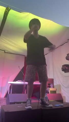The video begins with a full-body shot of a young man wearing a black t-shirt, leopard-print shorts, and black high-top sneakers. He is on a stage, standing on a speaker, with his right arm raised. He is lit by pink and yellow lights. The background shows a white tent, a table with a DJ setup, and some bottles of water. The camera then pans to show a person in the audience holding up a phone to record the performance. The scene then shifts to a blurry shot of a crowd of people outdoors at what appears to be a festival or concert. The sky is visible with clouds and a hint of sunset. The final shot shows a wider view of the crowd, with some people dancing and looking towards a stage in the distance, which is lit with colorful lights. A forested hill is visible behind the stage.