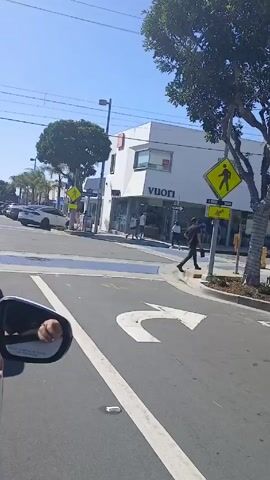 The video shows a street scene from the interior of a car stopped at a crosswalk. There is a Vuori store visible on the corner, and people are walking in the crosswalk. A white Rivian truck crosses in front of the car. The sky is blue and there are palm trees and other greenery along the street. Construction is taking place on a building further down the street.  The driver's arm is visible resting on the window. The cars are stopped at the crosswalk.