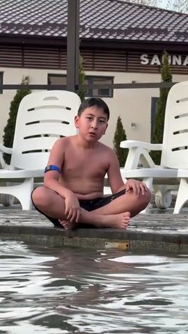 A young boy with wet hair is sitting cross-legged at the edge of a pool, wearing dark-colored swim trunks and a blue band on his left upper arm. He is looking forward with a neutral expression. Behind him are two white plastic lounge chairs and a building with a sign that says "SAUNA". The pool water is rippling, and a small portion of another person's head is visible in the lower right corner. The setting appears to be an outdoor pool area.