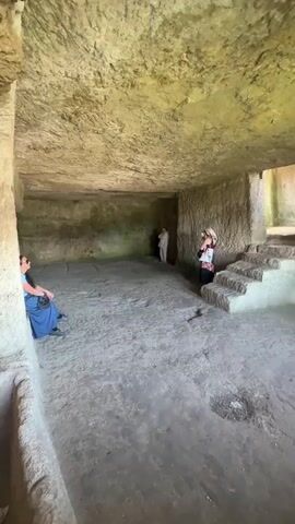 The video begins by panning across the interior of a cave-like dwelling. The walls and ceiling are rough-hewn stone, with a textured, yellowish-brown surface. The floor is also made of packed earth or stone. In the left of the frame, a person wearing a blue skirt and a dark top sits on the floor, facing away from the camera. In the background, a second person in a light-colored outfit stands near a doorway. To the right of the doorway, a set of rough-hewn stone stairs leads upwards. Further to the right, a third person, wearing a hat and a dark jacket over a colorful top, stands holding a camera, seemingly taking a photograph. The camera then moves to the right, focusing on the textured stone wall of the dwelling. The video then transitions to looking out from an opening in the cave. The opening frames a panoramic view of a lush green valley with rolling hills and dramatic, striped rock formations. The sky above is a clear, bright blue with a few wispy clouds. The perspective suggests the dwelling is situated high on a cliff or elevated landform, offering a commanding view of the landscape.
