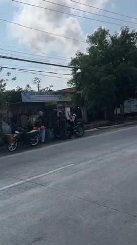 A group of people are gathered around several motorcycles parked on the side of a road next to a small building with a blue awning. The building appears to be a shop or stall. There are trees and power lines in the background, indicating an outdoor setting. The sky is partially cloudy.