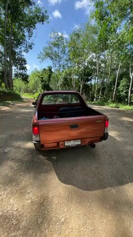A person is sitting in the driver's seat of a pickup truck. The truck is parked on a dirt road, surrounded by trees. The truck is orange, and it has a black toolbox and a red bag in the bed. The sky is blue with white clouds. The video is filmed from the perspective of someone in the bed of the truck, looking towards the cab. There is a sticker on the back window of the truck that says "BIQOKEE".
