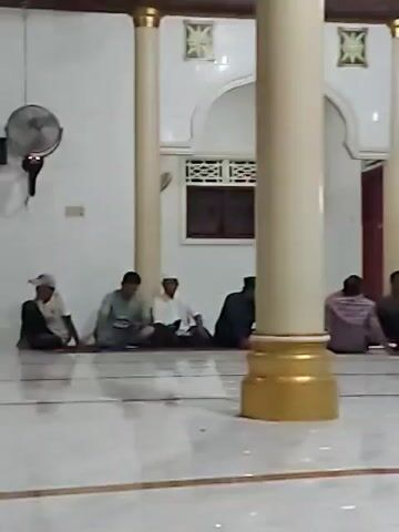 The video shows the interior of a mosque. Several men are sitting on the floor. The mosque has white tiled floors and walls, with tall, cream-colored pillars with gold bases. There are arched openings in the walls leading to other parts of the mosque. A fan is mounted on the wall, and there is artwork displayed. The overall lighting is moderate.