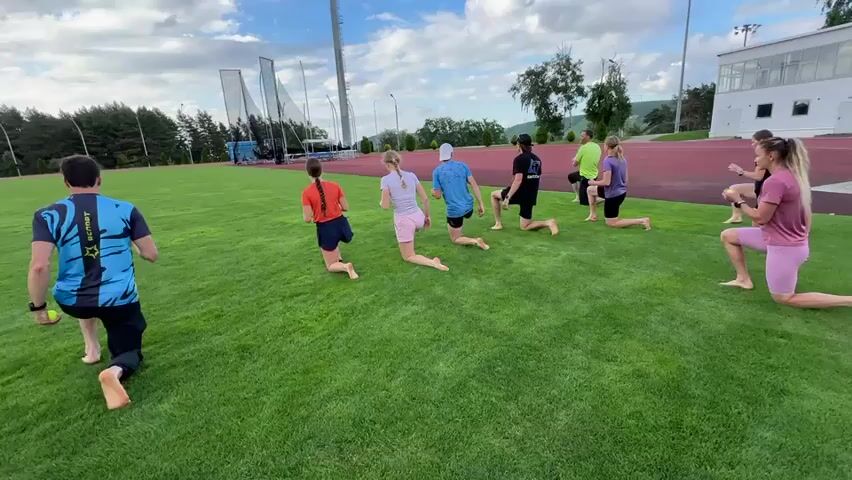 A group of at least eight people, mostly young women and one male coach, are exercising on a green grassy field. The coach, wearing a blue and black athletic shirt and black shorts, is in the foreground on the left, kneeling on his left knee and holding a yellow tennis ball in his right hand. The other participants are positioned in a line behind him, also in a lunging or kneeling position, with most of them also holding yellow tennis balls. The field appears to be part of an athletic facility, with a running track and stadium seating visible in the background, along with trees and a cloudy sky. The individuals are barefoot. The camera is positioned slightly behind and to the side of the coach, looking across the group.
