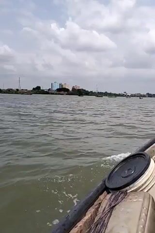 The video shows a boat moving across a body of water on a cloudy day. There are buildings on the shoreline in the distance, and a long bridge can be seen further away. The boat contains various items, including a solar panel and a white bucket with a black lid. There are two people in the boat. One is seated and appears to be steering. The other is a child who is eating something.