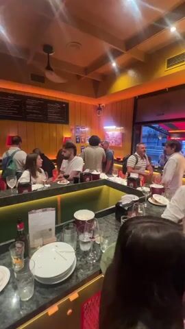 The video shows the interior of a bar or restaurant. People are seated at a bar and standing around. The decor includes yellow walls, wooden accents, and hanging lights. There are menus on the wall and bottles of alcohol behind the bar. On the bar in the foreground, there are plates with food, glasses, and a bottle of beer. In the background, staff members appear to be serving customers. The overall atmosphere seems lively and social.