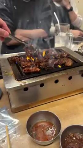 The video shows a small tabletop grill with pieces of marinated meat cooking on top. A pair of silver tongs flips the meat. Flames are visible below the grill, and smoke rises. There are two small metal bowls with sauces on the table next to the grill. People are seated around the table, but their faces are mostly out of view. One person wears a watch. There are also glasses with beverages and a small packet of food items on the table. The grill is placed on a wooden tabletop.