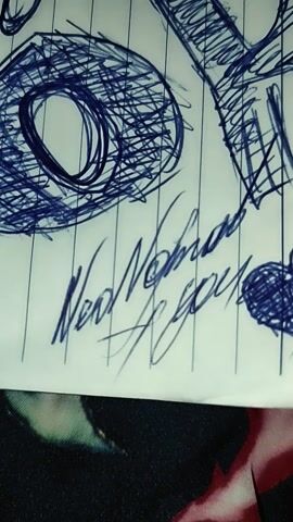 A close-up video shows a notebook with lined paper. The paper is covered in blue ink scribbles and drawings. There are a few distinct drawings including what appears to be a heart shape, and another shape that resembles a mask or eye. There are also some illegible words written in cursive. The notebook is lying on a surface that appears to be fabric with a floral pattern.