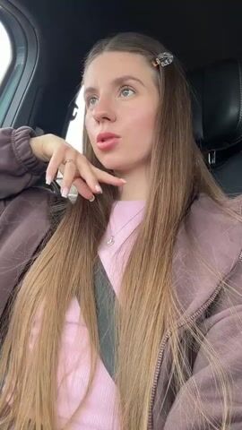 A young woman with long, straight brown hair, parted in the middle and adorned with two small, decorative hair clips, is seated in the passenger seat of a car. She is wearing a pink ribbed top layered under a dark brown or mauve zip-up hoodie. A silver necklace with a small pendant hangs around her neck. Her nails are manicured with a light polish.

Throughout the video, she poses for the camera, resting her chin on her hand, and shifts her gaze between looking directly at the camera and looking out the window. Her expressions vary from slightly pensive to a subtle, relaxed smile. The car's seatbelt is visible across her chest. The background outside the window suggests it's daytime, possibly overcast, with hints of trees or a highway barrier. Music is playing in the background. The scene depicts her enjoying a ride in the car.