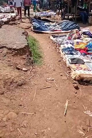 This video captures a bustling outdoor market scene, likely in Ngenyilel ward, Kenya. The ground is uneven and covered in dirt and debris, suggesting it's a well-worn path. The setting appears to be a market day, with numerous stalls set up along both sides of a central walkway.

The stalls display a wide array of goods, primarily clothing and fabrics. They are laid out on the ground, sometimes covered by tarps, showcasing colorful textiles, folded garments, and patterned materials. In the background, more permanent structures like market stalls or shops are visible, along with people milling about and engaging in commerce.

Several individuals are seen walking through the market. Two men in the foreground are walking away from the camera, one wearing a pink shirt and the other a white shirt. Further into the market, more people are visible, some appearing to be vendors and others customers. One person sits on a stool behind a stall, and others are seated or standing amongst the goods. A motorcycle is parked near a stall on the left side of the frame.

The sunlight suggests it is daytime, likely morning or afternoon, with bright lighting casting shadows. The presence of various fabrics, the layout of the stalls, and the interactions among people all point to a vibrant local marketplace. The uneven terrain and the exposed earth indicate a rural or less developed urban environment. There are no discernible text elements visible in the video. The overall atmosphere seems lively and active, typical of a community market.