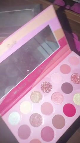 The video shows a close-up view of two open makeup palettes. The first palette is pink with a mirror and features various shades of pink, brown, and glitter. The brand "Rude" is visible on the top left of the palette. The second palette is black with a mirror and features shades of brown, pink, red, and gold. Both palettes are displayed on a wooden surface with other cosmetic items in the background.