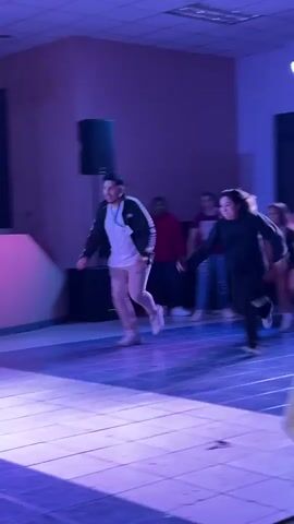 A group of people are on a stage with purple and blue lights. A young man in a white t-shirt and a black and white jacket is on the left. A woman in a black hoodie and black pants is on the right. She appears to be reaching out to the man, and then turns to the side and raises her arm. Another young man in a white t-shirt is standing behind them, holding a microphone. A screen with a logo is projected on the back wall. There is a small table with a bell on it on the right side of the stage. The people appear to be dancing or participating in a game.