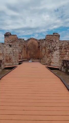 A low-angle, wide shot shows a curved wooden walkway extending toward ancient stone ruins under a partly cloudy sky. The walkway is made of horizontal planks, colored a warm, reddish-orange. The ruins are constructed of large, rough-hewn stone blocks, showing signs of age and decay. The central structure has a large archway, partially obscured by shadows, and flanking walls with broken buttresses and missing sections. The sky above is a vibrant blue with scattered white, fluffy clouds. The overall impression is one of historical significance and weathered grandeur.