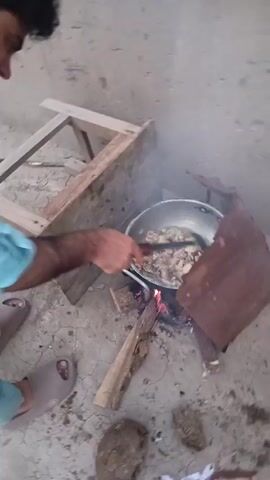 A young man in a light blue shalwar kameez is cooking outdoors. He is using a makeshift stove consisting of a metal container with a fire burning inside, fueled by wood. He is stirring something in a pot with a ladle. The scene appears to be a simple outdoor area with a dirt or concrete ground. In the background, there is a light-colored wall. The overall impression is of a basic, rural setting, likely in Kakki, Pakistan. The lighting suggests it is daytime. There is no visible text in the video. The man's focus is on the cooking, and there are no strong emotional cues visible. Debris, including scraps of fabric, is scattered on the ground around the cooking area.