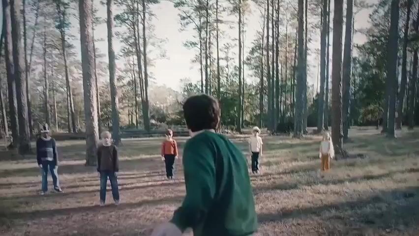 In a wooded area with tall trees and dry grass, a young boy with blonde hair runs away from the camera into the brush. He is wearing a brown jacket and blue jeans. Behind him, a man with dark hair, wearing a green sweater and blue jeans, is running towards the camera. In the distance, several other children are scattered throughout the woods, also running.
