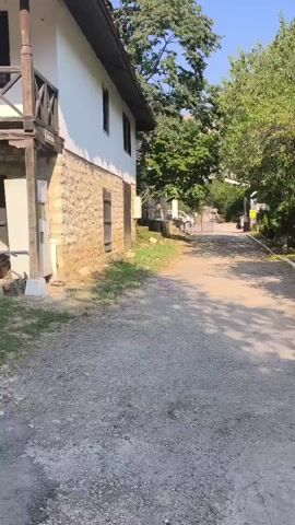 The video opens with a view of a traditional stone house with a wooden balcony. A wheelbarrow and some construction materials are visible next to the house. The camera then pans to show a person from behind, wearing a backpack, walking on a gravel path. Further on, two people are seen standing near a white board in a grassy area. The camera then focuses on a white SUV parked on the gravel path. The video continues to show the SUV and the traditional houses in the background, surrounded by trees.