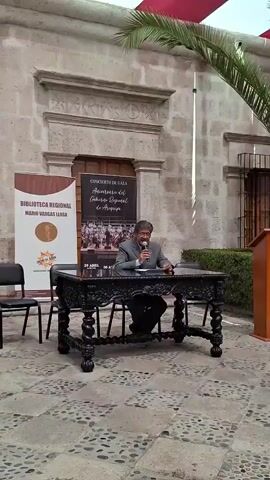 A man in a dark suit is seated at a large, ornate dark wooden table with spiral legs, speaking into a microphone. He is looking at papers on the table. Behind him are two banners. The left banner reads "BIBLIOTECA REGIONAL MARIO VARGAS LLOSA" and displays the logo of the "GOBIERNO REGIONAL AREQUIPA". The right banner announces a "CONCIERTO DE GALA Aniversario del Gobierno Regional de Arequipa" and shows dates "29 ABRIL" and "30 A..." (April), with an image of an orchestra. Two black chairs are visible, one next to the man, and another empty one further back. To the right of the table is a wooden podium. The setting is an outdoor courtyard or plaza with a patterned stone-paved ground and a historic stone building with carved details in the background. Green bushes are visible behind the table, and palm tree leaves are at the top right of the frame. The event appears to be a public address or announcement related to a cultural or governmental anniversary, likely in Arequipa, Peru, given the visible text on the banners. The time of day is bright and sunny, indicating clear weather.